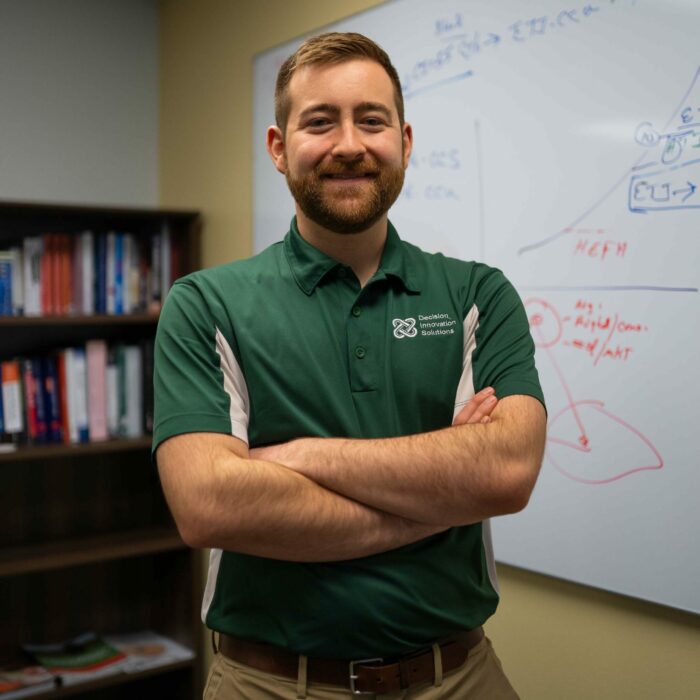 A bearded man with short hair stands confidently with folded arms, wearing a green polo shirt with a logo on the chest He is in an office setting with shelves of books and a whiteboard covered in diagrams and notes behind him